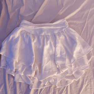 Vit volangkjol med lager på lager - Supersöt vit kortkjol med flera lager volanger som ger en flowy och luftig känsla och inbyggda shorts. Kjolen har bred linning och är tillverkad i ett lätt tyg, perfekt för varma dagar. Passar dig som gillar en romantisk och lekfull stil.