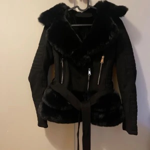 Svart pälsjacka med bälte och dragkedjor - Snygg svart pälsjacka med stora fuskpälsdetaljer på krage och nederkant. Jackan har markerade axlar, quiltade partier på ärmarna, två dragkedjefickor framtill och ett knytbälte i midjan. Perfekt för dig som vill sticka ut med en lyxig look.