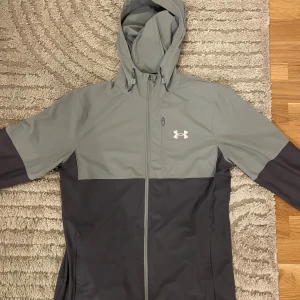 Grå och svart vindjacka Under Armour - Snygg tvåfärgad vindjacka från Under Armour i grått och svart. Jackan har huva, hel dragkedja framtill och en bröstficka med dragkedja. Perfekt för blåsiga dagar och sportiga outfits. Under Armour-logga på bröstet ger en sportig vibe.