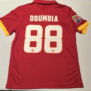 AS Roma Doumbia matchtröja Nike - AS Roma fotbollströja med Doumbia och nummer 88 på ryggen. Tröjan är röd med gula detaljer, officiellt Serie A-märke på ärmen och klubbmärke på bröstet. Tillverkad i Dri-FIT-material som andas. Klassisk pikékrage och korta ärmar. Replika, endast handtvätt gäller!