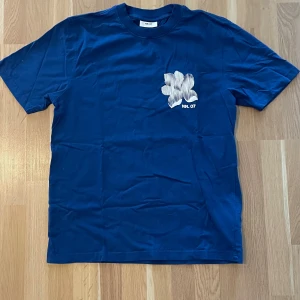 Blå t-shirt från NN.07 med tryck - Snygg marinblå t-shirt från NN.07 med vit blomtryck och logga på bröstet. Klassisk rund hals och korta ärmar. Perfekt för dig som gillar stilrena plagg med en twist. Materialet är mjuk bomull som känns skönt mot huden.