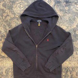 Mörkblå hoodie från Polo Ralph Lauren - Mörkblå hoodie från Polo Ralph Lauren med dragkedja och klassisk röd broderad logga på bröstet. Tröjan har huva, känguruficka och är tillverkad i mjuk bomull. Perfekt för en chill och stilren look. Hör av er vid minsta fundering!