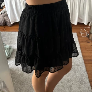 Svart prickig volangkjol från FB Sister - Supersöt svart kortkjol från FB Sister i storlek S. Kjolen har flera lager med volanger och ett prickigt mönster i tyget. Den har en elastisk midja som gör den bekväm och lätt att bära. Perfekt för dig som gillar en fin och trendig stil.