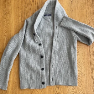 Grå ribbad kofta från Tommy Hilfiger - Str XS/S nästan oanvänd snygg grå kofta från Tommy Hilfiger i ribbstickad ull och kashmirmix. Koftan har lång ärm, v-ringning och svarta knappar framtill. Perfekt för dig som gillar stilrena och mjuka plagg med lite lyxig känsla. Passar dig som vill ha en klassisk look med modern touch.