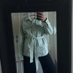 Mintgrön overshirt från Zara med bälte - Snygg mintgrön overshirt från Zara i jeansmaterial. Jackan har stora bröstfickor, knappar framtill och ett matchande bälte i midjan för en schysst siluett. Perfekt att slänga över en hoodie eller t-shirt. Långärmad och med klassisk krage.