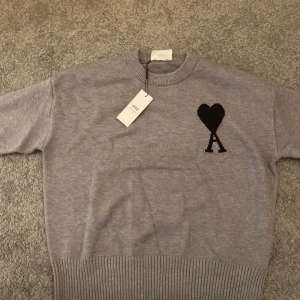 Grå stickad tröja från Ami Paris - Säljer en snygg grå stickad tröja från Ami Paris med svart hjärta och A-logga på bröstet. Tröjan har ribbade muddar vid hals, ärmslut och nederkant samt långärmad passform. Perfekt för dig som gillar stilrena detaljer och mjuka material.pris 📍kan diskuteras vid snabb affär!