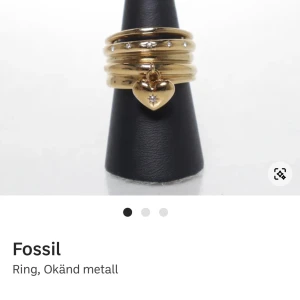 Guldfärgad ring med hjärthänge från Fossil - Snygg guldfärgad ring från Fossil med flera band i lager-på-lager look. Ringen har små glittriga stenar och ett hängande hjärta med en liten sten i mitten. Vattentålig. Köpt för 300:-