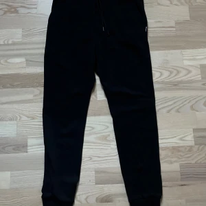 Svarta mjukisbyxor Jack & Jones - Svarta mjukisbyxor från Jack & Jones med elastisk midja och snörning. Byxorna har en klassisk, avslappnad passform och är tillverkade i mjukt material som passar perfekt för chill eller träning. Snygga muddar vid bensluten för en modern look.