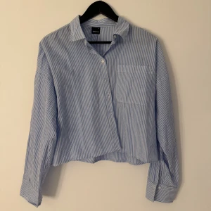 Blåvit randig skjorta från Gina Tricot - Snygg blå och vit randig skjorta från Gina Tricot med klassisk krage, bröstficka och knappar framtill. Skjortan har lång ärm och en lite croppad passform.
