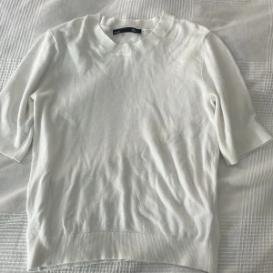 Vit stickad tröja från 157, S - Vit stickad tröja från 157 med ribbade muddar vid hals, ärmslut och nederkant. Tröjan har korta ärmar och en klassisk rund halsringning. Perfekt för dig som gillar en clean och enkel stil. Har använd några gånger men fortfarande i bra stick.