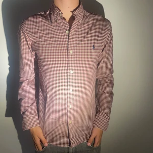 Rutig skjorta från Ralph Lauren - Säljer en snygg rutig skjorta från Ralph Lauren i slim fit. Skjortan har en klassisk krage och är långärmad med knappar hela vägen ner. Den är i en stilren kombination av röd och vit, perfekt för både vardag och fest. Märket är broderat på bröstet för en extra touch av stil.