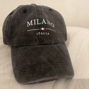 Svart keps Milano Italia - Snygg svart keps med vit text 'MILANO ITALIA' och en liten italiensk flagga framtill. Klassisk böjd skärm och justerbart spänne bak. Tillverkad i bomullstyg med tvättad look som ger en cool vintagekänsla.