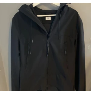 CP company jacka - Säljer en svart softshell jacka från C.P. Company med ikoniska glasögon i huvan. Jackan har dragkedja framtill, två bröstfickor med dragkedja och justerbar huva med snörning. Skyddar bra mot kyla speciellt med tröja under. Skriv för fler bilder!