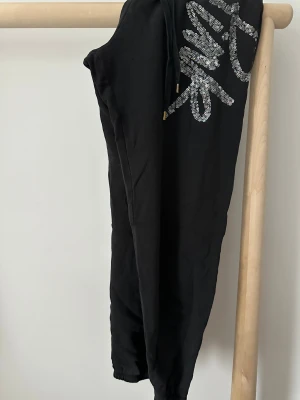 Svarta mjukisbyxor med paljetter - Svarta mjukisbyxor från Victoria's Secret PINK med glittrande silverpaljetter på ena benet. Byxorna har snörning i midjan och muddar vid bensluten för en skön och avslappnad passform. 