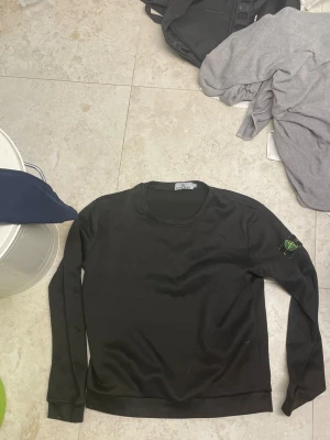Svart långärmad tröja Stone Island - Svart långärmad tröja från Stone Island med klassisk rund hals och ikonisk patch på vänster ärm. Tröjan är i mjukt bomullsmaterial och har en clean, stilren look som funkar till jeans eller cargo. Perfekt för dig som gillar streetwear och vill ha något enkelt men ändå med edge.