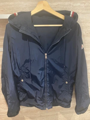 Mörkblå vindjacka från Moncler - Snygg mörkblå vindjacka från Moncler med huva och dragkedja framtill. Jackan har två fickor med tryckknappar, snörning i huvan och Moncler-logga på ärmen. Perfekt för blåsiga dagar och har coola rödvita detaljer på huvan. Skriv vid frågor