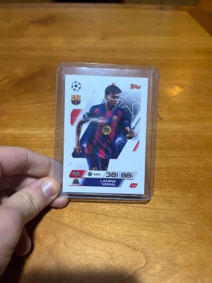 Blå-röd randig FC Barcelona väst - Ett officiellt Topps Match Attax-kort med Lamine Yamal i Barcelonas hemmatröja. Kortet har en ren, modern design med UEFA-Champions-League-tema och visar spelarens värden: 38 DEF och 88 ATT. Kortnumret är 113, och bilden framhäver Yamal i aktion som en av Barcelonas unga, lovande offensiva spelare.