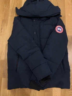 Mörkblå dunjacka från Canada Goose - Säljer en mörkblå dunjacka från Canada Goose size S-M med huva och klassisk logga på ärmen. Jackan har quiltad design, långa ärmar och stängs med både dragkedja och knappar. Perfekt för kalla vinterdagar och har en clean, stilren look.