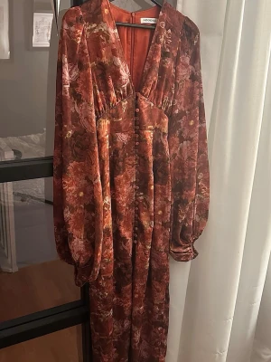 Blommig långklänning från Adoore - Långklänning från Adoore i varma rödbruna toner med blommigt mönster. Klänningen har v-ringning, långa puffärmar och dekorativa knappar framtill. Materialet är mjukt och följsamt, perfekt inför jul!