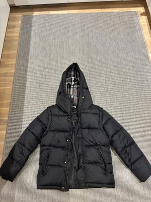 Svart pufferjacka från Burberry - Svart pufferjacka från Burberry med klassiskt rutigt foder i grått, vitt och rött. Jackan har huva, dragkedja och tryckknappar framtill samt flera fickor. Yttermaterialet är polyester och fodret är mjukt och värmande. Perfekt för kalla dagar.