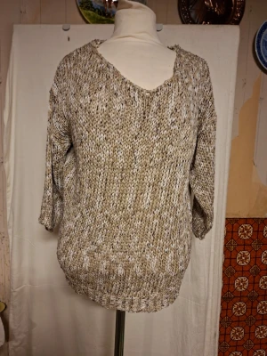 Beige stickad tröja Leidiró Firenze - Snygg beige stickad tröja från Leidiró Firenze med v-ringad hals och trekvarts ärmar. Framsidan är grovstickad i beige och vitt, medan baksidan är slät och satinliknande. Perfekt för dig som gillar unika detaljer och skön passform.