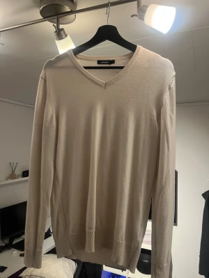 Beige v-ringad tröja J.Lindeberg M - Snygg beige v-ringad tröja från J.Lindeberg i storlek M. Tröjan har lång ärm, diskret JL-logo broderad på bröstet och ribbade muddar vid ärmslut och nederkant. Perfekt för dig som gillar stilrena och klassiska plagg.