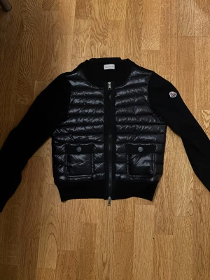 Svart Cardigan från Moncler - Svart Cardigan från Moncler med quiltad framsida, två fickor med knappstängning och dragkedja. Jackan har en stilren look med Moncler-logga på ärmen och är tillverkad i polyester och dun. Perfekt för dig som gillar exklusiva märken och snygg design.
