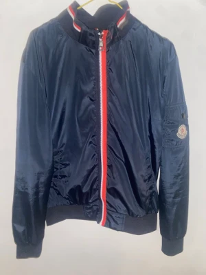 Moncler marinblå bomberjacka - Säljer en snygg marinblå bomberjacka från Moncler med röd och vit dragkedja samt ribbad krage med ränder. Jackan har lång ärm, Moncler-logga på ärmen och glansigt polyestermaterial. Perfekt för dig som vill ha en clean och sportig look.