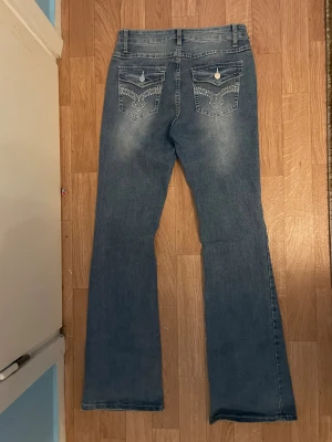 Blå bootcut jeans med dekorerade fickor - Snygga blå bootcut jeans med coola detaljer på bakfickorna, inklusive silverfärgade knappar och broderade mönster. Jeansen har klassisk femficksdesign och är tillverkade i mjukt denimtyg. Perfekt för dig som gillar en avslappnad och trendig stil.