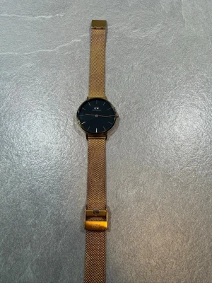 Guld klocka från Daniel Wellington - Snygg klocka från Daniel Wellington med mesharmband i guld och svart urtavla. Stilren och minimalistisk design med tunna guldiga visare och index. Låset är markerat med DW och armbandet är i rostfritt stål. Perfekt accessoar för att lyfta din outfit.