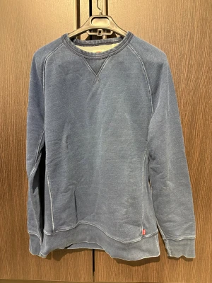 Blå sweatshirt från Levi's - Säljer en chill blå sweatshirt från Levi's med klassisk rund hals och synliga sömmar. Tröjan har lång ärm och en liten röd Levi's-tag vid nederkanten. Perfekt för dig som gillar enkel och stilren streetwear.