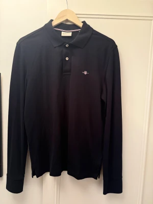 Svart långärmad pikétröja från GANT - Svart långärmad pikétröja från GANT med klassisk krage och två vita knappar framtill. Tröjan har broderad GANT-logga på bröstet och är gjord i mjuk bomull. Perfekt för dig som gillar stilrena och tidlösa plagg.