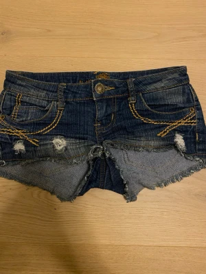 Blå jeansshorts från Almost Famous - Snygga blå jeansshorts från Almost Famous med låg midja och råa, fransiga kanter. Shortsen har detaljer, gul dekorationssöm och klassiska fem fickor. Perfekt för sommarens chill dagar och festivalhäng.