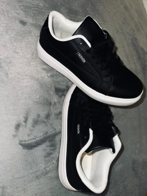 Svarta sneakers med vit sula - Svarta sneakers från Fashion med vit platt sula och vit insida. Skorna har snörning och en rund tå. Ovandelen är i syntetmaterial och designen är stilren med diskreta sömmar. Perfekt för dig som gillar klassiska och enkla sneakers.