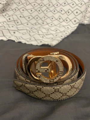 Beige mönstrat bälte från G - Snyggt bälte från Gu i beige och brunt med ett klassiskt G-mönster och en stor, rund, guldig metallspänne med G-logga. Bältet har en lyxig känsla och är tillverkat i skinn med tryckt mönster. Perfekt för att lyfta din outfit med en exklusiv touch.