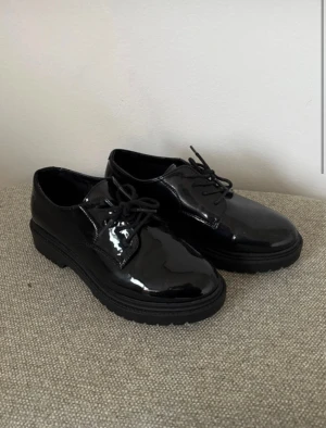Svarta lackloafers med snörning - Snygga svarta loafers med glansig lackyta och klassisk snörning framtill. Skorna har rund tå och platt sula, vilket ger en stilren och modern look. Perfekta för dig som vill ha en dressad vibe med lite edge.