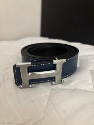 Blått läder bälte med silver spänne - Jag säljer ett blått stiligt bälte med ett silver spänne. Bältet är 125 cm och kan vändas in och ut från blått till svart.