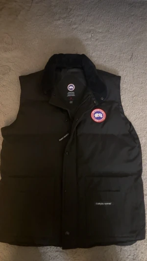 Svart pufferväst från Canada Goose - Svart pufferväst från Canada Goose med klassisk röd och vit logotyp på bröstet. Västen har hög krage, två stora fickor framtill och dragkedja med tryckknappar. Perfekt för kalla dagar och har en clean, stilren look.