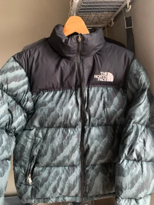 The North Face pufferjacka M - Säljer en fet pufferjacka från The North Face i storlek M. Jackan har mörkgrön och svart färg med coolt mönster, hög krage och dragkedja framtill. Perfekt för kalla dagar och har två fickor med dragkedja. Loggan syns tydligt på bröstet. Har sytts en gång på högra underarmen men syns knte mycket (se bild 4). Skriv för fler bilder eller andra frågor!