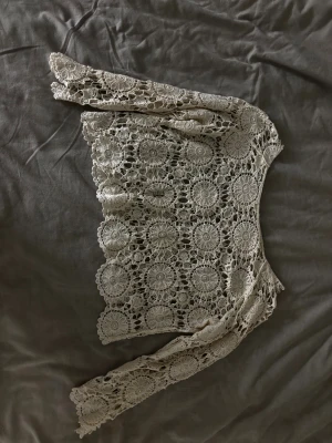 Beige virkad bolerotopp med spets - Säljer en beige bolerotopp med lång ärm, helt virkad i ett detaljerat spetsmönster med cirkulära blommor. Toppen är kortare i modellen och har en bohemisk vibe med öppet, luftigt material. Perfekt för att lägga över en klänning eller linne. Den är designad som kortare i armarna.