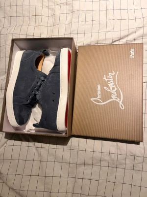 Christian Louboutin blå mockasneakers - Snygga sneakers från Christian Louboutin i mörkblå mocka med vit platt sula och ikonisk röd undersida. Skorna har rund tå, snörning och beige innersula med logga. Perfekt för dig som vill sticka ut med exklusiv stil och detaljer.