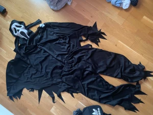 Svart Scream-dräkt med mask, storlek 164 - Säljer en svart Scream-kostym i storlek 164 med tillhörande vit mask. Dräkten har taggiga kanter på ärmar och ben för en skräckig look. Materialet är 100% polyester och passar perfekt till halloween eller temafest. Masken är klassisk och lätt att bära. Säljer även några magväskor. Kom med pris