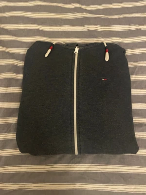 Hilfiger Denim Zip Hoodie  - Hoodie från Hilfiger Denim med grått foder i huvan och vita snören med röda och svarta detaljer. Tröjan har hel dragkedja, två fickor fram och klassisk Hilfiger-logga på bröstet. Perfekt för chill dagar och enkel att matcha med jeans.