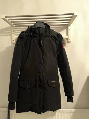 Svart Canada Goose parkas med päls - Svart parkas från Canada Goose med ikonisk röd logotyp på ärmen, stor huva med beige pälsdetalj och praktiska fickor framtill. Jackan har dragkedja och knappar, samt justerbar midja för snygg passform. Perfekt för kalla vinterdagar.