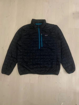 Svart pufferjacka Patagonia XL - Quiltad svart pufferjacka från Patagonia i storlek XL. Jackan har blå detaljer, halv dragkedja framtill och en bröstficka med dragkedja. Lätt och smidig modell med hög krage, perfekt för kyliga dagar. I perfekt skick