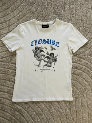 Vit t-shirt med ängeltryck från Closure - Vit t-shirt från Closure London med blå text och stort tryck av två änglar och duvor på framsidan. Klassisk rund hals och korta ärmar. Skön och lätt i bomull, perfekt för en chill och trendig look. Endast använd 2 gånger 