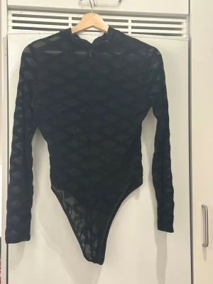 Svart mesh bodysuit med diamantmönster - Säljer en svart mesh bodysuit med lång ärm och högt skuren nederdel. Toppen har ett genomskinligt material med diamantformat mönster och en liten krage. Perfekt för att skapa en edgy look med lager på lager.