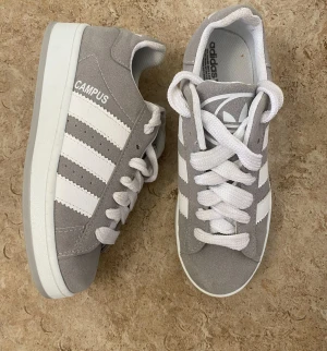 Grå Adidas Campus sneakers - Snygga Adidas Campus sneakers i ljusgrå mocka med klassiska vita ränder och vit sula. Skorna har rund tå, platt sula och vita snören. Perfekta för dig som gillar en clean och stilren look med retrovibbar.