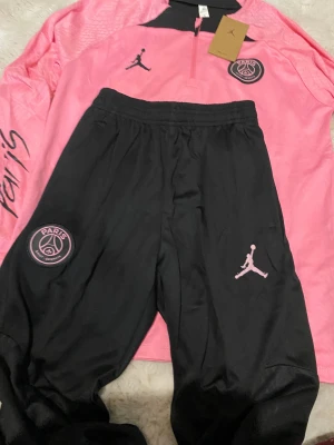 Psg track suit jordans  - Bilderna visar endast ett rosa och svart PSG x Jordan träningsset med långärmad tröja och byxor, men inga skor finns med.