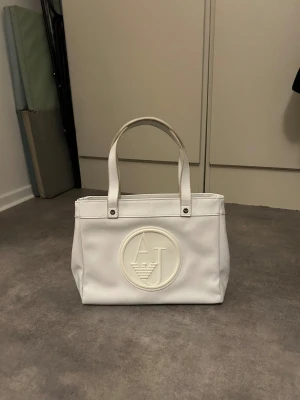 Vit handväska från Armani Jeans - Snygg vit handväska från Armani Jeans med AJ-logga i relief på framsidan. Väskan har två rejäla handtag, flera fack med dragkedja och är tillverkad i blankt syntetmaterial. Perfekt storlek för att få plats med allt du behöver och stilren design.